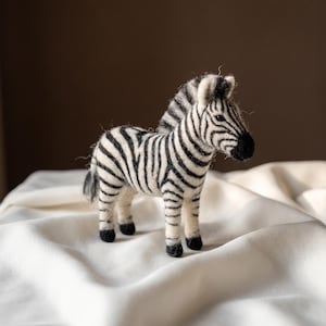 May include: A needle-felted zebra figurine featuring a white body with distinct black stripes, a black mane, a black tail, and black hooves. The zebra is standing in a profile position, showcasing its textured wool construction and detailed facial features.