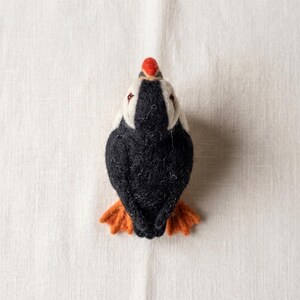 May include: A needle-felted puffin figurine featuring a black back and wings, a white face, and a bright orange beak. The bird has small, textured orange feet extending from the bottom, with a soft, fuzzy appearance throughout the entire body.