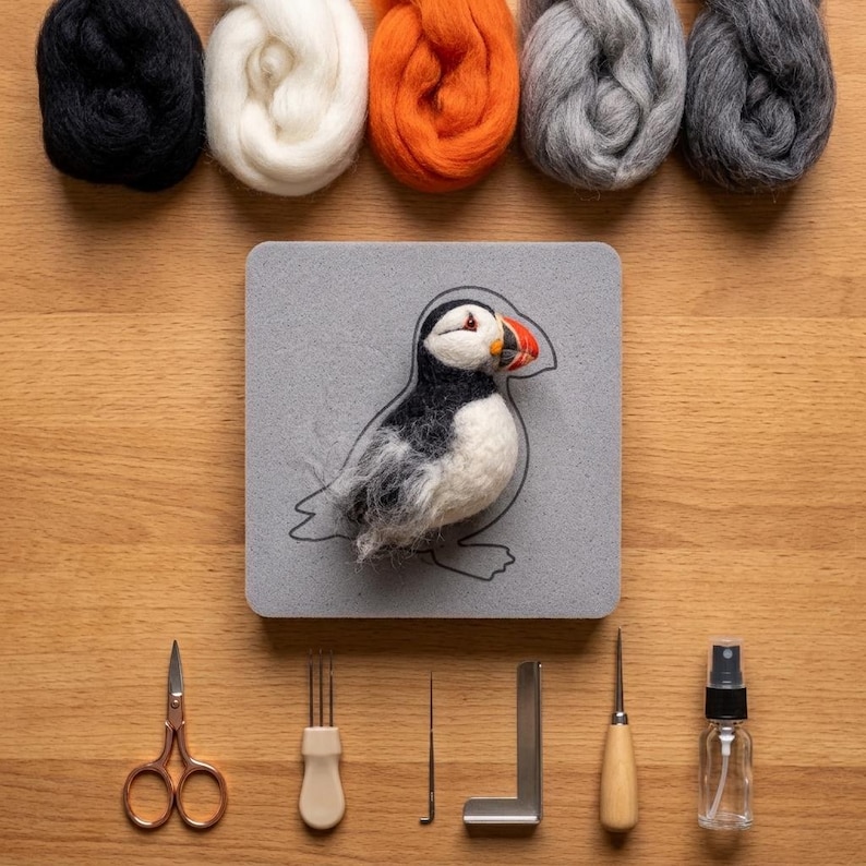 May include: A needle felting kit featuring a puffin figure on a gray foam pad, five bundles of wool roving in black, white, orange, light gray, and dark gray, copper-toned scissors, a multi-needle tool, a single needle, a metal square, and a spray bottle.