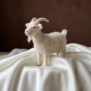 May include: A needle-felted white goat figurine with a textured wool finish, curved tan horns, and a small beard. The goat stands in a profile position, showcasing detailed facial features, pointed ears, and a short tail, crafted with soft fibres.