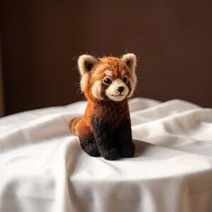 May include: A needle-felted red panda figurine with a textured, fuzzy coat in shades of reddish-brown, black, and cream. The small animal has pointed ears, dark eyes, a black nose, and a white muzzle, sitting in an upright, alert posture.