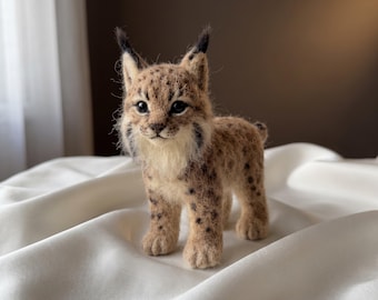 May include: A needle-felted lynx figurine featuring a tan coat with dark brown spots, a cream-colored chest, and black-tipped ears. The small animal sculpture has dark, glossy eyes and a detailed face with whiskers, standing in a realistic pose.