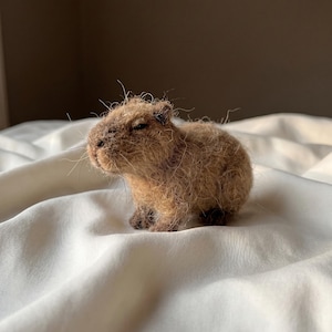 Peut inclure: Une figurine de capybara en laine feutrée à l'aiguille, composée de fibres de laine brun clair texturées. La petite sculpture animale présente un corps arrondi, un museau émoussé, de petites oreilles sombres et des pattes brun foncé.