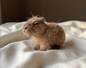 Op de afbeelding: Een naaldgevilte capybara-figuur gemaakt van getextureerde lichtbruine wolvezels. Het kleine dierensculptuur heeft een rond lichaam, een stompe snuit, kleine donkere oren en donkerbruine voeten, waarbij losse vezels een pluizig uiterlijk creëren.