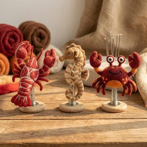 Könnte beinhalten: Drei nadelgefilzte Meerestierfiguren auf Metallfedern und Holzsockeln. Ein roter Hummer, ein beiges Seepferdchen mit weißen Streifen und eine dunkelrote Krabbe mit orangefarbenen Punkten sind in einer Reihe angeordnet, jeweils mit zwei kleinen weißen Augen.
