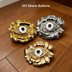 May include: Three decorative shank buttons in gold and silver, shaped like flowers with an eye design. The buttons have a white center with a black dot and are embellished with small rhinestones. The text "DIY Shank Buttons" is at the top.