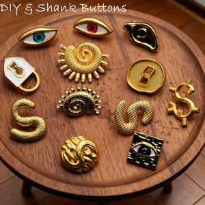 Puede incluir: Una colección de botones dorados con vástago en varios diseños, incluyendo ojos, un candado, un signo de dólar y la letra "S". Los botones están dispuestos sobre una bandeja de madera, con el texto "DIY & Shank Buttons" en la parte superior.
