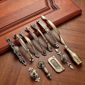May include: A collection of antique brass door handles and pulls in various designs. The hardware includes ornate handles, a bottle-shaped pull, and a recessed pull, all with a vintage aesthetic. The items are arranged on a wooden surface.