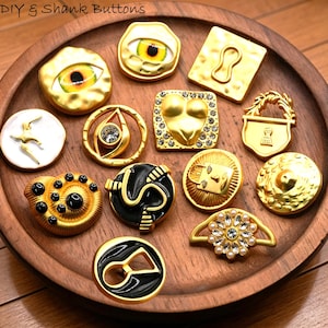May include: A collection of gold-colored shank buttons with various designs, including an eye, a keyhole, a lion's face, and a floral design. The buttons are displayed on a wooden tray. The text "DIY & Shank Buttons" is visible.