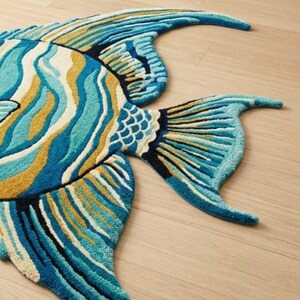 May include: A colourful, fish-shaped rug in shades of blue, teal, and gold. The rug has a detailed design with textured patterns, resembling scales and fins. The rug is placed on a light-coloured wooden floor.