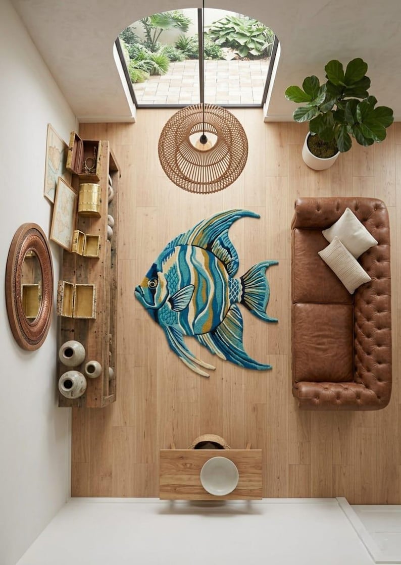 May include: A decorative fish-shaped rug in shades of blue, yellow, and white, placed on a wooden floor. The room features a brown leather sofa, a wooden shelf, and a woven light fixture. The rug's dimensions are approximately 180 cm.
