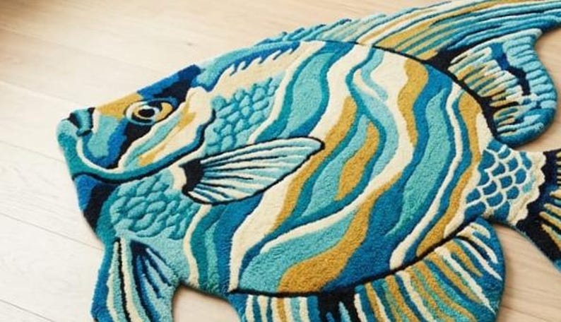 May include: A colourful fish-shaped rug with a vibrant design. The rug features shades of blue, teal, yellow, and white, creating a wave-like pattern across the body. The fish has detailed fins and a textured surface, adding depth to the decorative floor covering.