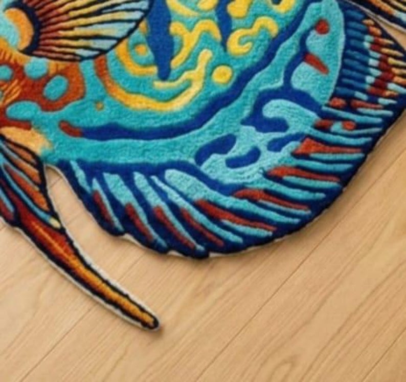 Op de afbeelding: Een kleurrijk, visvormig tapijt met een gedetailleerd ontwerp. Het tapijt heeft tinten blauw, turquoise, geel, oranje en rood. Het tapijt ligt op een lichtgekleurde houten vloer.