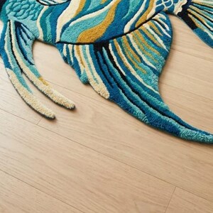 May include: A vibrant, fish-shaped rug with a detailed design. The rug features shades of blue, teal, and gold, with a textured, tufted appearance. The fish has fins and a tail, creating a whimsical and decorative floor covering.