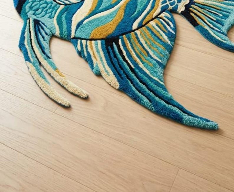 May include: A vibrant, fish-shaped rug with a detailed design. The rug features shades of blue, teal, and gold, with a textured, tufted appearance. The fish has fins and a tail, creating a whimsical and decorative floor covering.