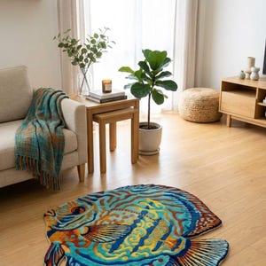 Op de afbeelding: Een kleurrijk, visvormig vloerkleed met een gedetailleerd ontwerp in blauw, geel en oranje. Het kleed ligt op een houten vloer in een woonkamer en voegt een decoratief element toe aan de ruimte. Afmetingen: ongeveer 150 cm.