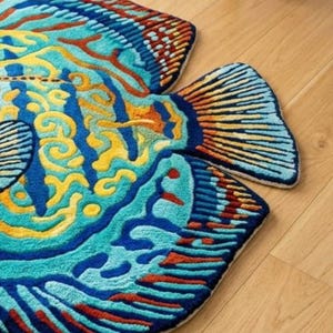 Op de afbeelding: Een kleurrijk, visvormig tapijt met een gedetailleerd ontwerp. Het tapijt heeft tinten blauw, teal, geel en oranje, met ingewikkelde patronen. Het tapijt ligt op een houten vloer en toont zijn levendige kleuren en unieke vorm.