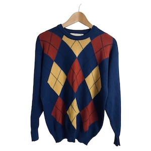 Può includere: Un maglione a maniche lunghe con girocollo di colore blu, caratterizzato da un motivo a rombi nei toni del giallo senape e del rosso ruggine. Il capo in maglia presenta collo, polsini e orlo a coste ed è appeso a una gruccia in legno.