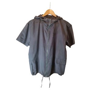 Puede incluir: Chaqueta gris oscuro de manga corta con capucha, colgada de una percha de madera. La chaqueta tiene una cremallera completa, dos bolsillos delanteros y un cordón en la parte inferior. La tela parece ser ligera.