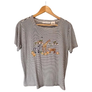 Puede incluir: Camiseta a rayas con cuello redondo y manga corta. La camiseta presenta un diseño bordado de una cebra, una jirafa y un leopardo, con pequeños detalles florales. La camiseta está colgada de una percha de madera.
