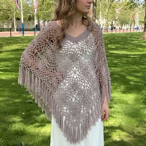 May include: A mauve crochet poncho featuring an intricate floral pattern with an openwork design and a V-neckline. The garment is finished with long, straight fringe along the bottom edges, creating a pointed silhouette that drapes over the shoulders.