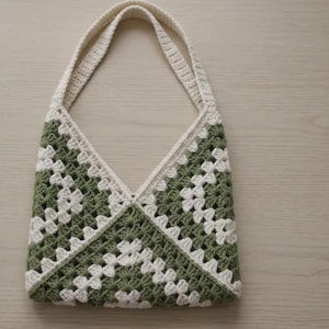 May include: A handmade crochet tote bag featuring a classic granny square pattern in alternating sage green and off-white yarn. The bag has a triangular V-shaped top opening and two integrated shoulder straps crafted from the same crocheted yarn.