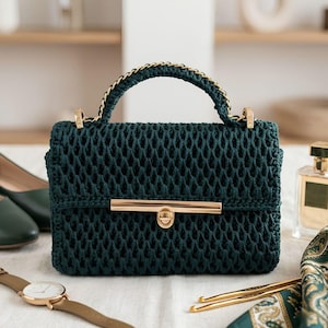 Peut inclure: Un sac à main en crochet vert foncé présentant une forme rectangulaire structurée, une poignée supérieure avec des détails de chaîne dorée et une fermeture à barre métallique dorée sur le rabat avant au motif en losanges texturé.