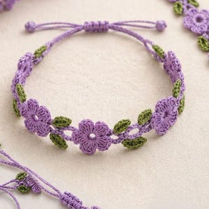 May include: A crochet bracelet featuring a series of lavender purple flowers and small green leaves connected by thin purple thread. The bracelet includes an adjustable sliding knot closure with two dangling ends finished with small purple knots.