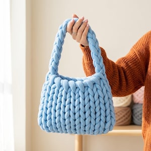 Può includere: Una borsa a mano azzurra realizzata con filato a maglia spesso e voluminoso. La borsa presenta un corpo rettangolare strutturato con un motivo a treccia verticale e un unico manico intrecciato integrato che si inarca sopra la borsa.
