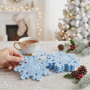 Könnte beinhalten: Ein Set aus hellblauen, gehäkelten Schneeflocken-Untersetzern mit filigranen Mustern. Ein Untersetzer liegt unter einer weiß-gold gesprenkelten Keramiktasse, während ein Stapel identischer Untersetzer daneben auf einer weißen Holzoberfläche liegt.