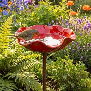 May include: A red, petal-shaped bird bath with a rusty brown stand, filled with water. A small bird with orange and brown feathers drinks from the water. The bird bath is surrounded by lush green foliage and colorful flowers, creating a vibrant garden scene.