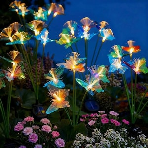 Pode incluir: Uma cena de jardim com luzes solares de fibra ótica em forma de borboletas e flores. As luzes são em várias cores, incluindo azul, verde e laranja, e são colocadas entre flores cor-de-rosa e brancas. O fundo é azul escuro.