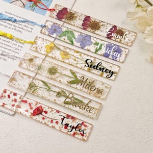 Puede incluir: Una colección de marcapáginas rectangulares de resina, cada uno con flores secas y copos dorados. Nombres como "Emma", "Mickey", "Sidney", "Milena", "Brooke" y "Taylor" están escritos en los marcapáginas. Cada marcapáginas tiene una borla.