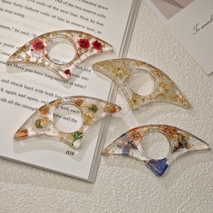May include: Four clear resin thumb page holders, each with a unique design. One features red flowers, another gold flakes, a third with small flowers, and the last with blue flowers. The page holders are on a book.