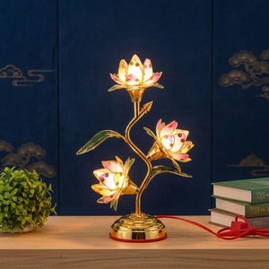 Könnte beinhalten: Eine goldfarbene Metalllampe mit drei beleuchteten Lotusblüten mit rosa und weißen Blütenblättern. Die Lampe hat einen schlanken, geschwungenen Stiel mit grünen Blättern und einen runden Sockel, verbunden mit einem roten Stromkabel mit Schalter.
