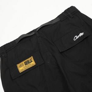 Puede incluir: Pantalones cargo negros con m&uacute;ltiples bolsillos y cintur&oacute;n. Los pantalones tienen un parche amarillo y negro que dice "Guerillaz" y un logotipo blanco en el bolsillo. Los pantalones est&aacute;n hechos de un material duradero.
