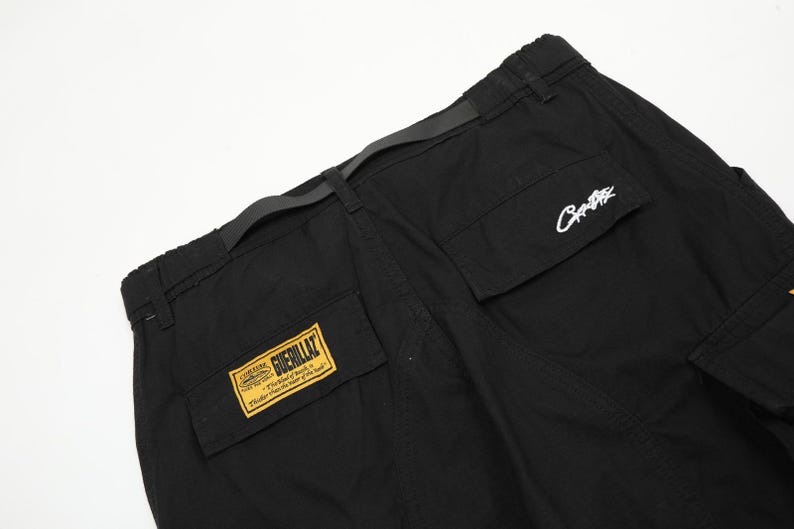 Puede incluir: Pantalones cargo negros con m&uacute;ltiples bolsillos y cintur&oacute;n. Los pantalones tienen un parche amarillo y negro que dice "Guerillaz" y un logotipo blanco en el bolsillo. Los pantalones est&aacute;n hechos de un material duradero.