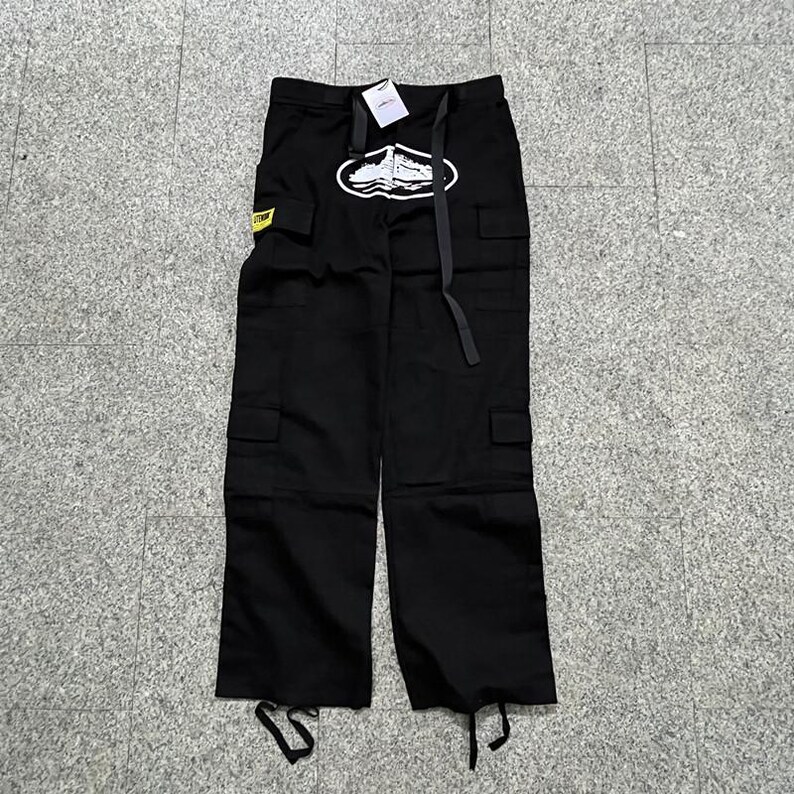 Puede incluir: Pantalones cargo negros con m&uacute;ltiples bolsillos y un gr&aacute;fico blanco en la pierna izquierda. Los pantalones tienen cintura con cord&oacute;n y una etiqueta amarilla con texto. El gr&aacute;fico tiene forma ovalada con un dise&ntilde;o de barco.