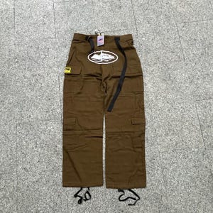 Puede incluir: Pantalones cargo marrones con m&uacute;ltiples bolsillos y un cintur&oacute;n negro. Los pantalones presentan un dise&ntilde;o ovalado blanco con un gr&aacute;fico de barco en la parte delantera. Una peque&ntilde;a etiqueta amarilla es visible en el lado izquierdo.