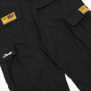 Puede incluir: Pantalones cargo negros con m&uacute;ltiples bolsillos y parches rectangulares amarillos. Un parche dice "Guerillaz". Los pantalones tienen un corte holgado y est&aacute;n hechos de una tela duradera. Un logotipo blanco est&aacute; en uno de los bolsillos.