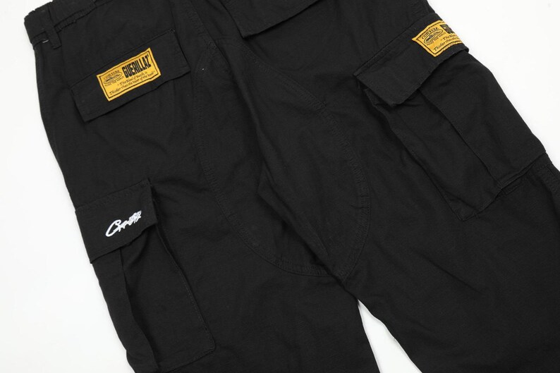 Puede incluir: Pantalones cargo negros con m&uacute;ltiples bolsillos y parches rectangulares amarillos. Un parche dice "Guerillaz". Los pantalones tienen un corte holgado y est&aacute;n hechos de una tela duradera. Un logotipo blanco est&aacute; en uno de los bolsillos.