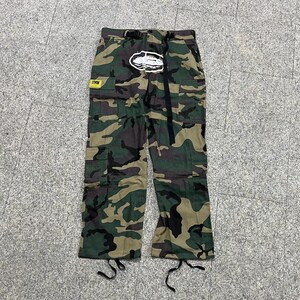 Puede incluir: Pantalones cargo de camuflaje con m&uacute;ltiples bolsillos, un cintur&oacute;n negro y un logotipo blanco en la parte delantera. Los pantalones presentan una mezcla de patrones verdes, marrones y negros. Una etiqueta amarilla es visible en el bolsillo izquierdo.
