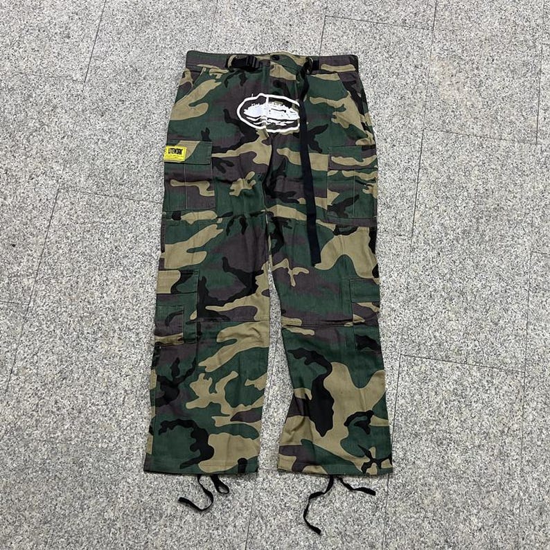 Puede incluir: Pantalones cargo de camuflaje con m&uacute;ltiples bolsillos, un cintur&oacute;n negro y un logotipo blanco en la parte delantera. Los pantalones presentan una mezcla de patrones verdes, marrones y negros. Una etiqueta amarilla es visible en el bolsillo izquierdo.