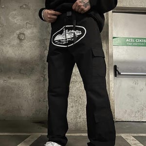 Puede incluir: Pantalones cargo negros con un dise&ntilde;o gr&aacute;fico blanco en la parte delantera. Los pantalones tienen m&uacute;ltiples bolsillos y un cintur&oacute;n. La persona lleva zapatillas blancas y una sudadera con capucha negra.