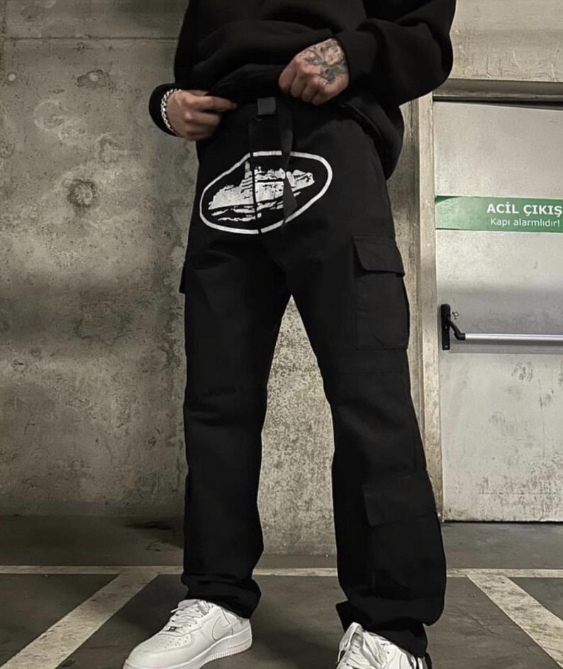 Puede incluir: Pantalones cargo negros con un dise&ntilde;o gr&aacute;fico blanco en la parte delantera. Los pantalones tienen m&uacute;ltiples bolsillos y un cintur&oacute;n. La persona lleva zapatillas blancas y una sudadera con capucha negra.