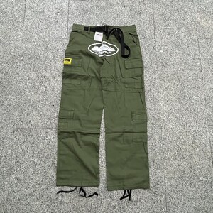 Puede incluir: Pantalones cargo verde oliva con m&uacute;ltiples bolsillos y cintur&oacute;n negro. Un gr&aacute;fico blanco est&aacute; centrado en la parte delantera y una etiqueta amarilla es visible en el bolsillo izquierdo. Los pantalones tienen un corte relajado y est&aacute;n hechos de tela duradera.