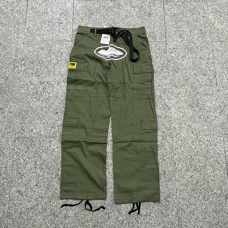 Puede incluir: Pantalones cargo verde oliva con m&uacute;ltiples bolsillos y cintur&oacute;n negro. Un gr&aacute;fico blanco est&aacute; centrado en la parte delantera y una etiqueta amarilla es visible en el bolsillo izquierdo. Los pantalones tienen un corte relajado y est&aacute;n hechos de tela duradera.