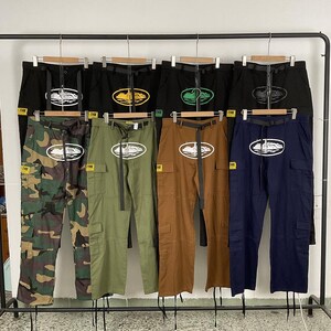 Puede incluir: Una variedad de pantalones cargo en negro, camuflaje, verde oliva, marr&oacute;n y azul marino, cada uno con un dise&ntilde;o de logotipo &uacute;nico. Los pantalones se exhiben en una percha, mostrando sus diferentes colores y estilos. Cada par tiene un cintur&oacute;n negro.