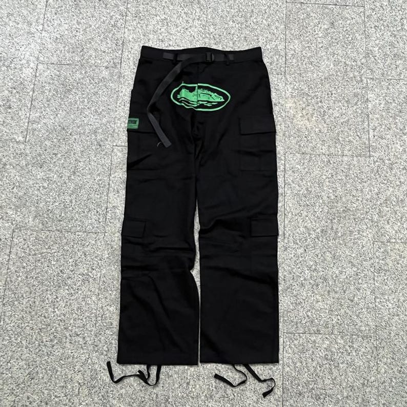 Puede incluir: Pantalones cargo negros con m&uacute;ltiples bolsillos y cintur&oacute;n. Un gr&aacute;fico ovalado verde con un dise&ntilde;o de zapato est&aacute; en la parte delantera. Un peque&ntilde;o parche cuadrado verde est&aacute; en el lado izquierdo. Los pantalones tienen cordones en la parte inferior.