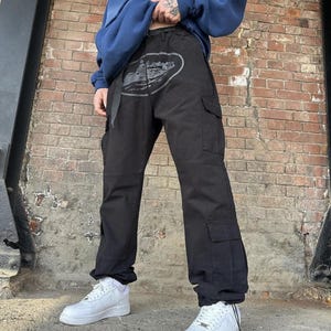 Puede incluir: Pantalones cargo negros con un dise&ntilde;o gr&aacute;fico gris en la parte delantera, con m&uacute;ltiples bolsillos y un cintur&oacute;n. Los pantalones se combinan con zapatillas blancas y una sudadera azul. El fondo es una pared de ladrillos.